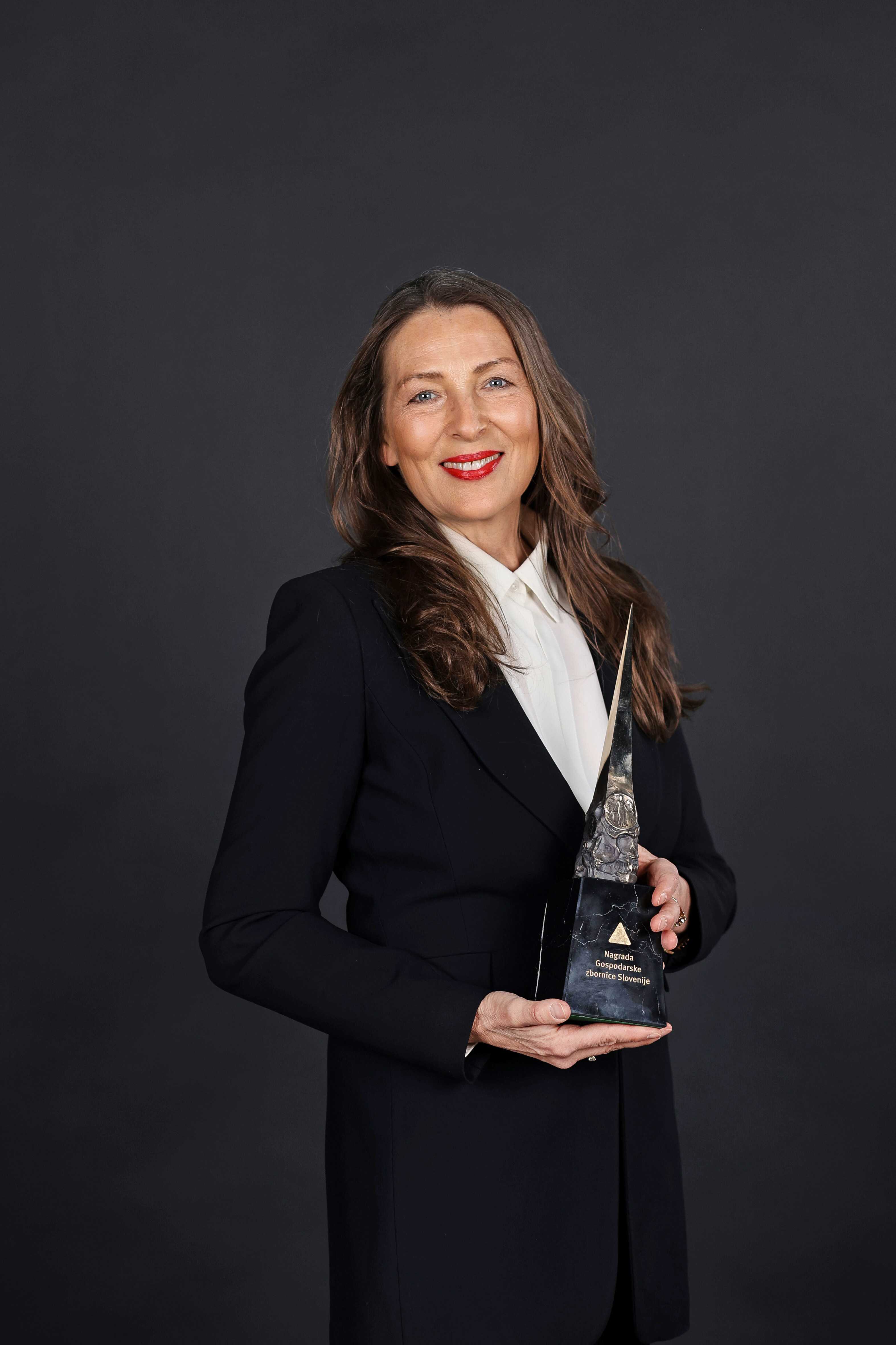 A professional portrait of Estera Brajak, dressed in a black blazer and white blouse, holding the Gospodarska zbornica Slovenije award. She has long, wavy brown hair and a confident smile, standing against a dark, neutral background. The award features a sharp, metallic design with an engraved plaque.